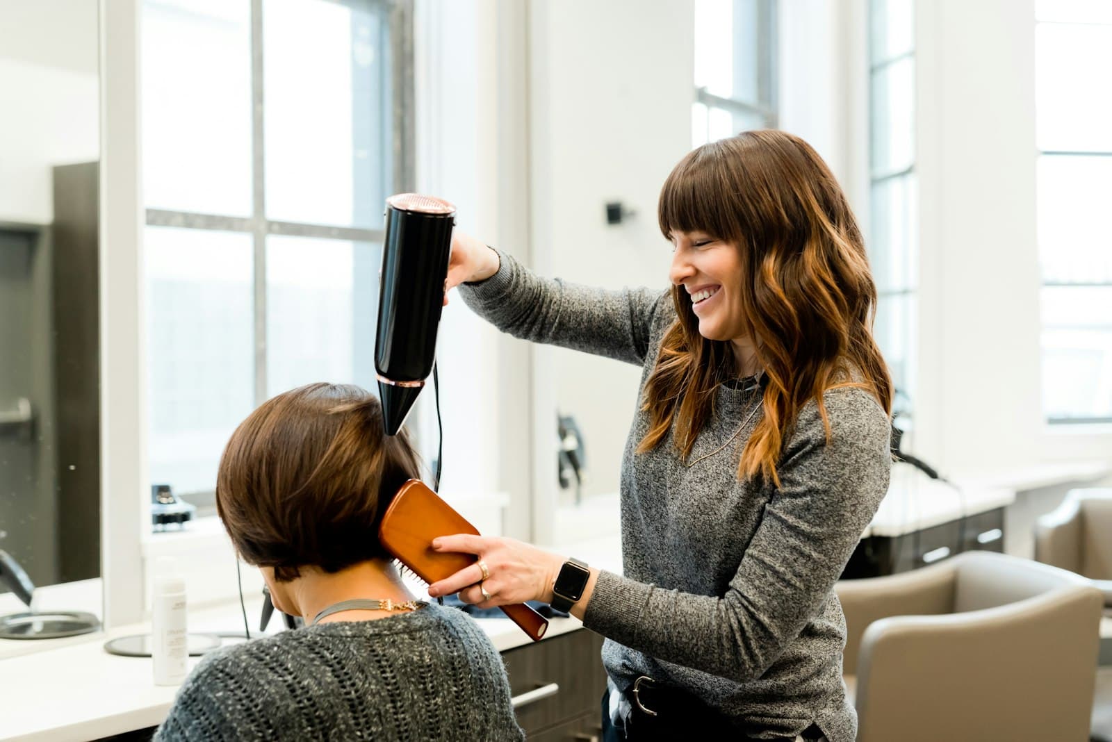 A stylist smiling while blow-drying a client's hair in a bright salon